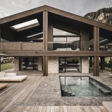 Vista laterale della facciata dell'Chalet Gardena di Cortina Hospitality, con dettagli in pietra e legno, salotto esterno, piscina incastonata e design che richiama le maestose montagne circostanti.