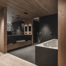 Camera padronale con bagno in legno, doppio lavabo, vasca da bagno e elemento di pietra all'Chalet Gardena di Cortina Hospitality, un'oasi di eleganza e materiali autentici.