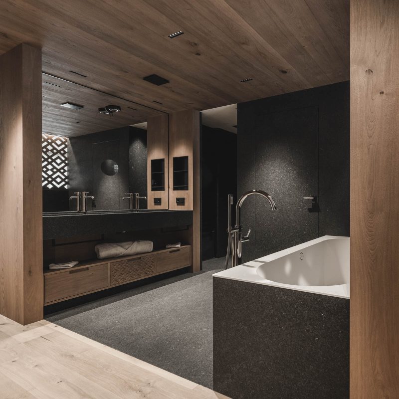 Camera padronale con bagno in legno, doppio lavabo, vasca da bagno e elemento di pietra all'Chalet Gardena di Cortina Hospitality, un'oasi di eleganza e materiali autentici.