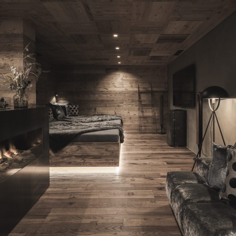 Camera da letto principale con caminetto e design raffinato nell'Chalet Seceda di Cortina Hospitality, finestre che offrono una vista incantevole del bosco.