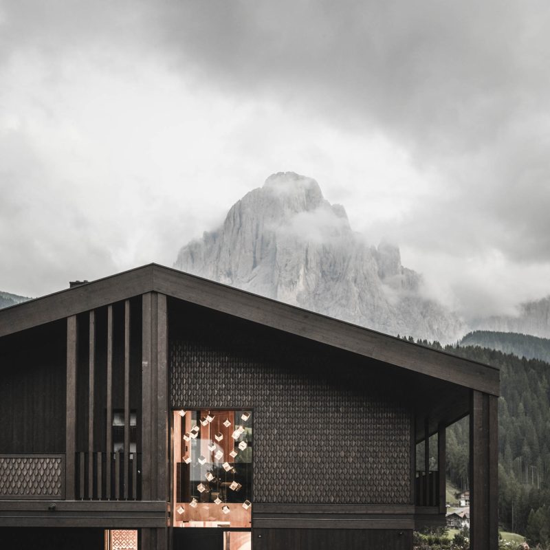 Vista esterna dell'Chalet Gardena di Cortina Hospitality, con dettagli delle pareti in legno e grandi finestre illuminate, creando una sinfonia di luce e design.