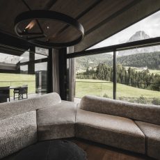 Angolo del divano con finestre panoramiche e terrazze esterne visibili all'Chalet Gardena di Cortina Hospitality, dove il design vetro elimina le barriere tra spazi interni ed esterni.