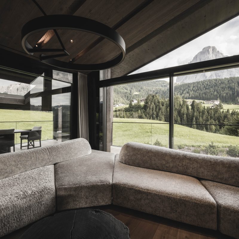 Angolo del divano con finestre panoramiche e terrazze esterne visibili all'Chalet Gardena di Cortina Hospitality, dove il design vetro elimina le barriere tra spazi interni ed esterni.