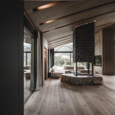Living in legno con caminetto a 360 gradi, pietra dolomitica e finestre panoramiche al mansardato dell'Chalet Gardena di Cortina Hospitality, offrendo un'esperienza immersiva nella natura.