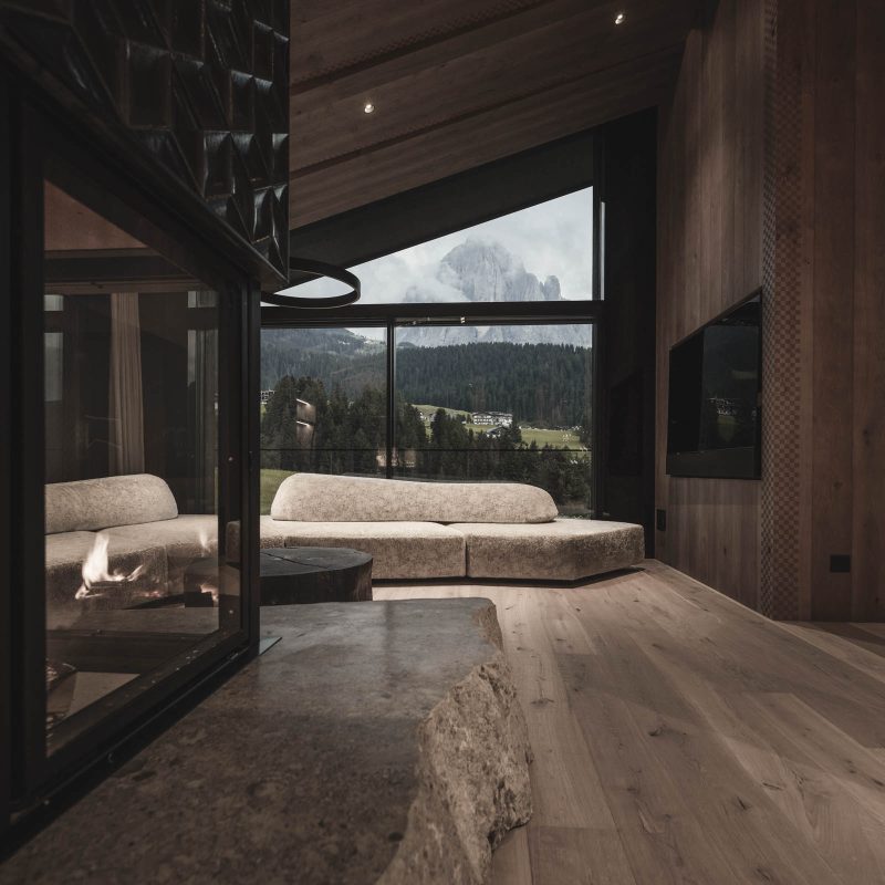 Living al tramonto con il calore del caminetto acceso all'Chalet Gardena di Cortina Hospitality, creando una scena magica e avvolgente.