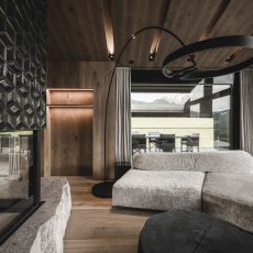 Divano ispirato alle montagne, caminetto in vetro su grande pietra e terrazza panoramica all'Chalet Gardena di Cortina Hospitality, rappresentando una connessione armoniosa con la natura circostante.