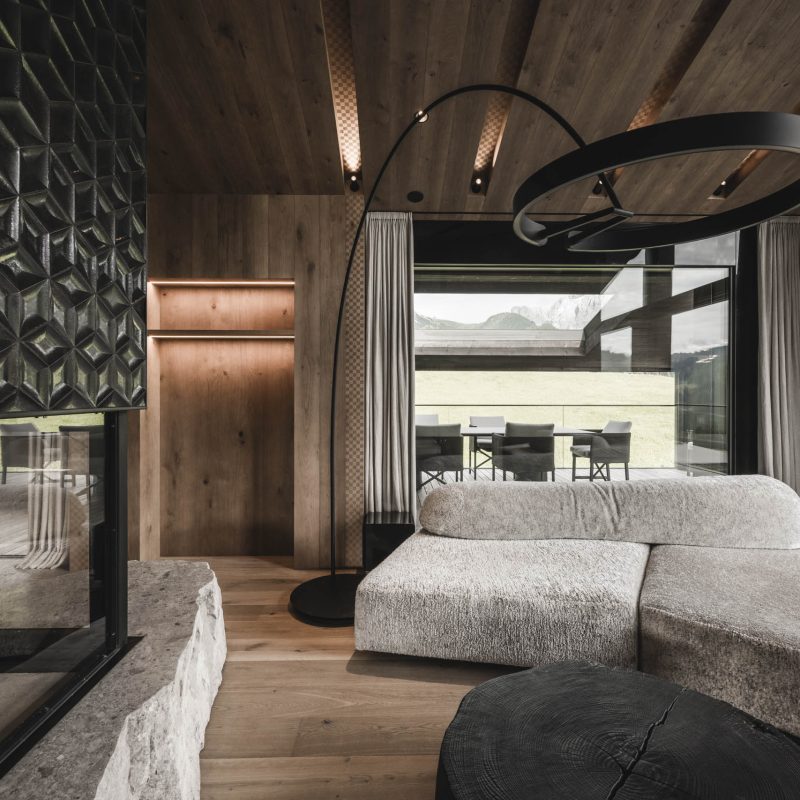 Divano ispirato alle montagne, caminetto in vetro su grande pietra e terrazza panoramica all'Chalet Gardena di Cortina Hospitality, rappresentando una connessione armoniosa con la natura circostante.